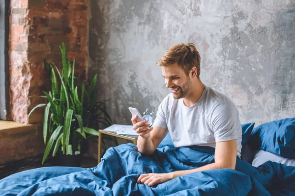 Lächelnder junger Mann checkt Smartphone zu Hause im Bett — Stockfoto