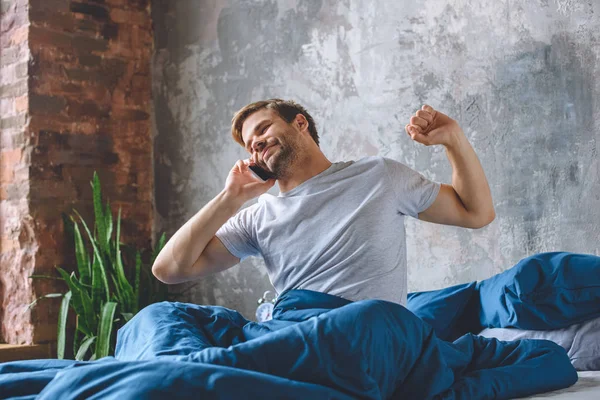 Young man talking on smartphone and stretching in bed during morning time at home — Stock Photo