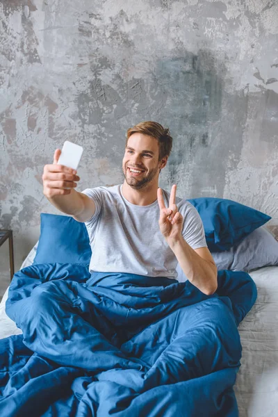 Joven feliz haciendo signo de paz y tomar selfie en el teléfono inteligente en la cama en casa - foto de stock