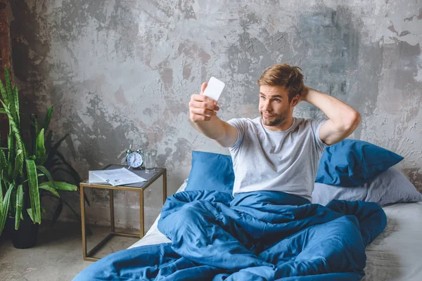 Bel giovanotto scattare selfie su smartphone a letto a casa — Foto stock