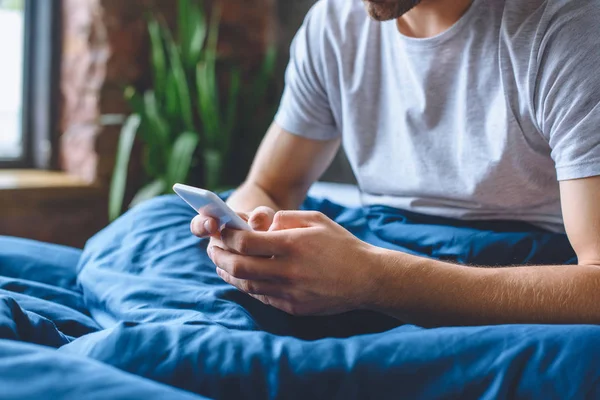 Частичный вид молодого человека, использующего смартфон в постели дома — стоковое фото