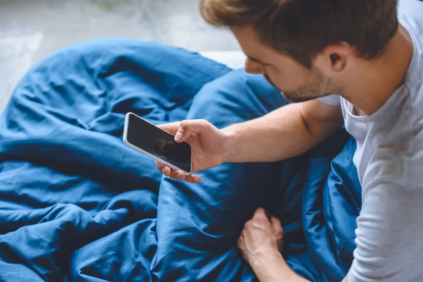 Enfoque selectivo de hombre joven en la cama con teléfono inteligente con pantalla en blanco - foto de stock