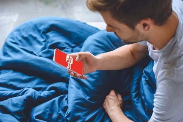 Messa a fuoco selettiva del giovane a letto utilizzando smartphone con youtube sullo schermo — Foto stock