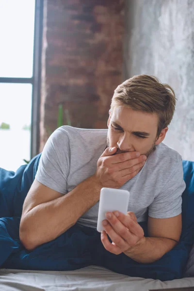 Young man yawning and checking smartphone in bed during morning time at home — Stock Photo