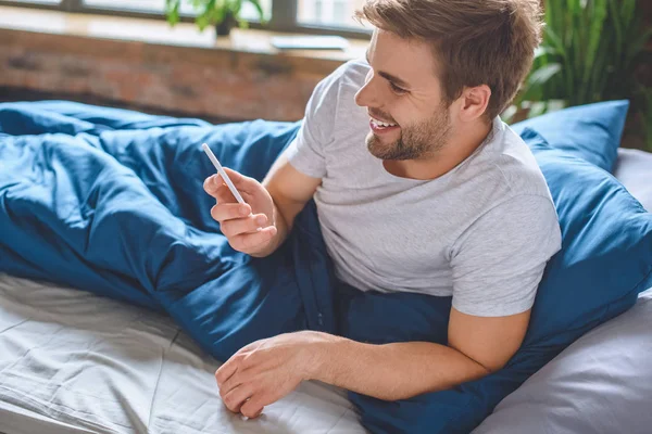 Sonriente joven comprobar teléfono inteligente en la cama en casa - foto de stock