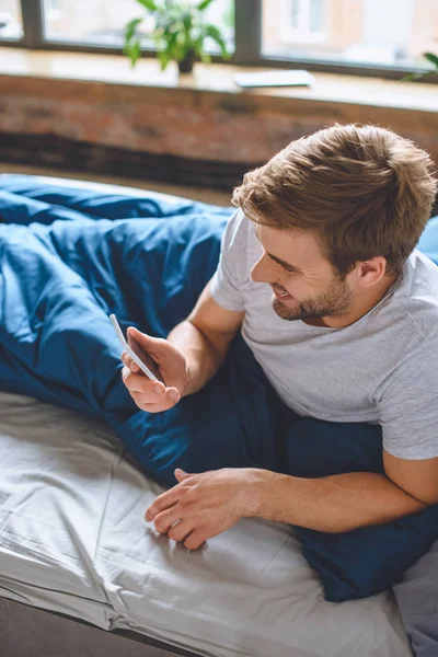 Vue grand angle de jeune homme souriant vérifier smartphone dans le lit à la maison — Photo de stock