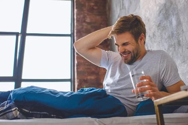 Giovane con mal di testa che tiene un bicchiere d'acqua a letto a casa — Foto stock