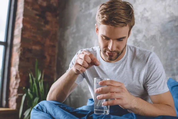 Young man putting pill in glass of water in bed at home — Stock Photo