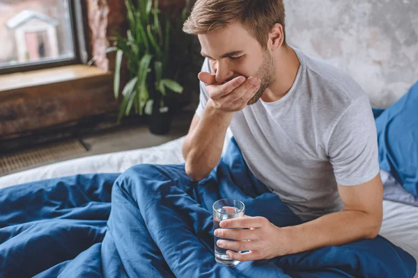 Selektiver Fokus eines jungen Mannes, der Tabletten nimmt und zu Hause ein Glas Wasser im Bett hält — Stockfoto