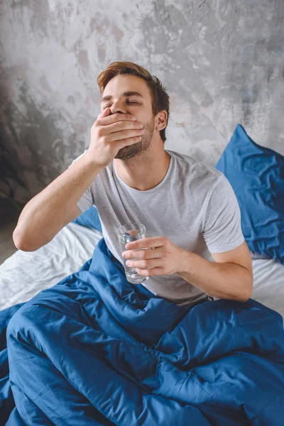 Blick aus der Vogelperspektive auf einen jungen Mann mit Kater, der Tabletten nimmt und zu Hause ein Glas Wasser im Bett hält — Stockfoto