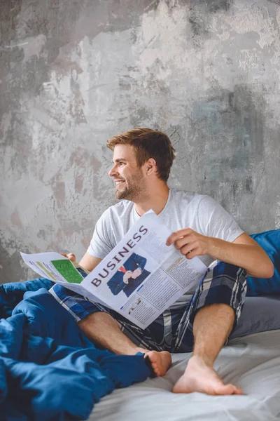 Усміхнений молодий чоловік читає бізнес-газету в ліжку в ранковий час вдома — стокове фото