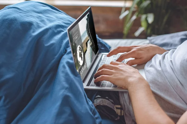 Imagen recortada del hombre en la cama usando el ordenador portátil con el sitio web de reserva en la pantalla - foto de stock