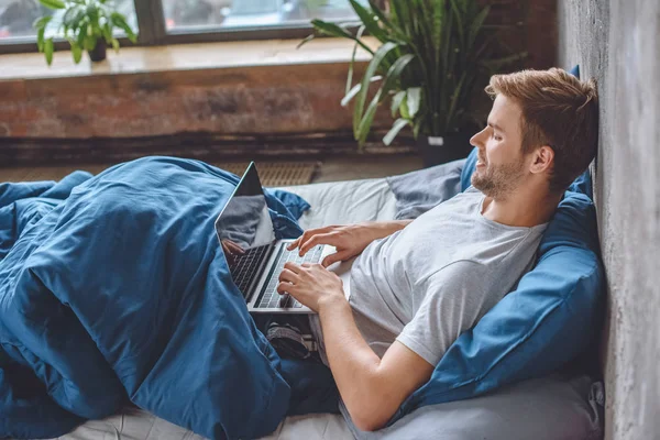 Smiling man in bed using laptop with booking website on screen — Stock Photo