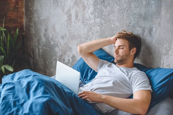 Расстроен молодой человек, использующий ноутбук в постели дома — стоковое фото