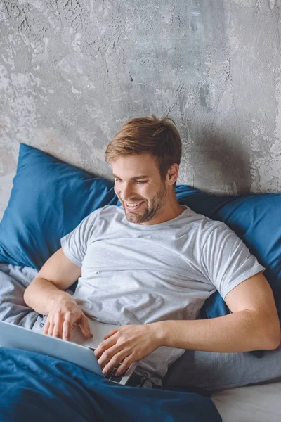 Heureux bel homme en utilisant un ordinateur portable dans le lit à la maison — Photo de stock