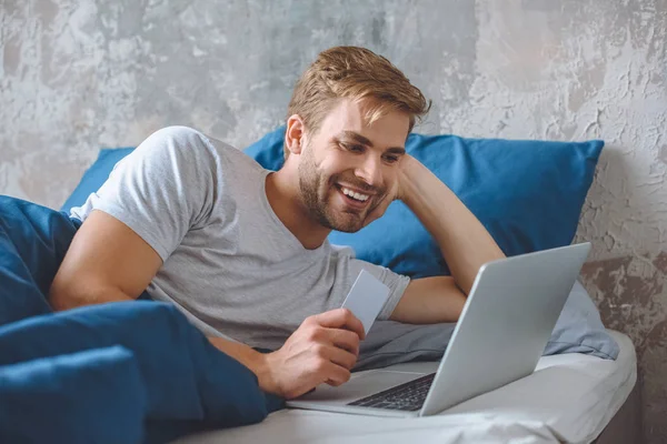 Sonriente joven en la cama con tarjeta de crédito haciendo compras en línea en el ordenador portátil - foto de stock