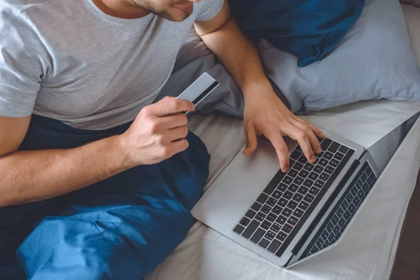 Обрезанное изображение человека в постели с кредитной картой, делающей онлайн покупки на ноутбуке — стоковое фото