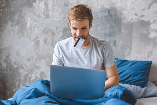 Jeune homme au lit avec carte de crédit dans la bouche faire des achats en ligne sur ordinateur portable — Photo de stock
