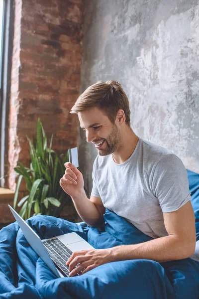 Heureux jeune homme au lit avec carte de crédit faire des achats en ligne sur ordinateur portable — Photo de stock