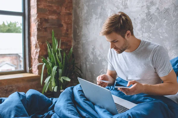 Jovem na cama com cartão de crédito e smartphone fazendo compras on-line no laptop — Fotografia de Stock