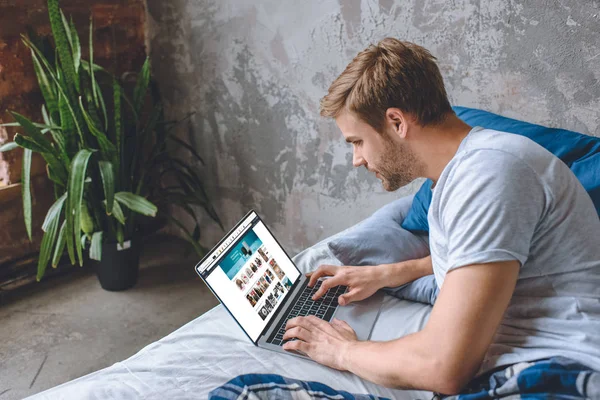 Joven en la cama usando el ordenador portátil con el sitio web de Amazon en la pantalla - foto de stock