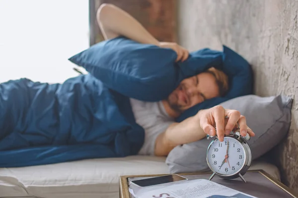 Angry young man in bed closing ear by pillow and turning off alarm clock during morning time at home — Stock Photo