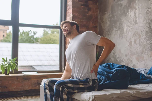 Молодий чоловік з болем у хребті просто прокидається і сидить на ліжку вдома — стокове фото