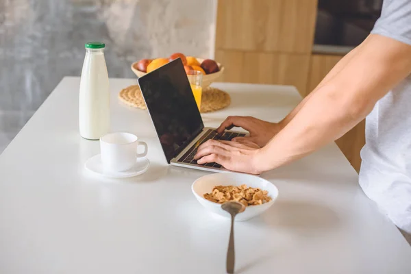 Image recadrée de jeune homme en utilisant un ordinateur portable à la table de cuisine avec petit déjeuner — Photo de stock