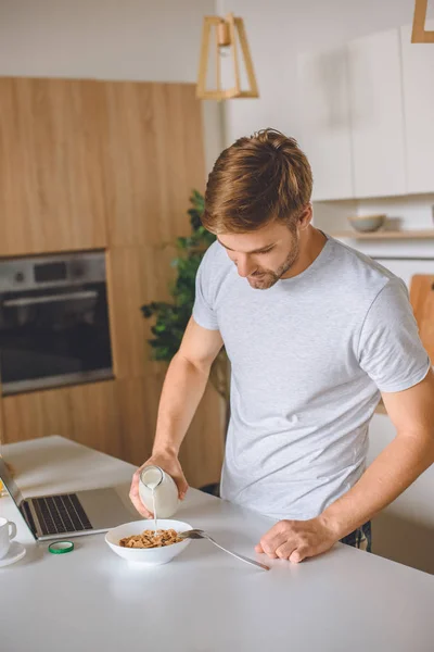 Young man pouring milk into plate with flakes at kitchen table with laptop — Stock Photo