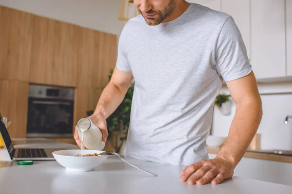 Imagem cortada do homem derramando leite em placa com flocos na mesa da cozinha com laptop — Fotografia de Stock