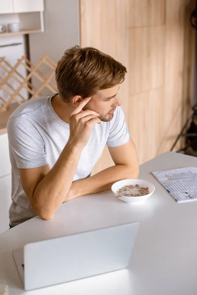 Vue grand angle de réfléchi jeune homme assis à la table de cuisine avec ordinateur portable et petit déjeuner — Photo de stock