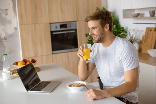 Homem sorridente bebendo suco fresco à mesa com prato de flocos e laptop na cozinha — Fotografia de Stock