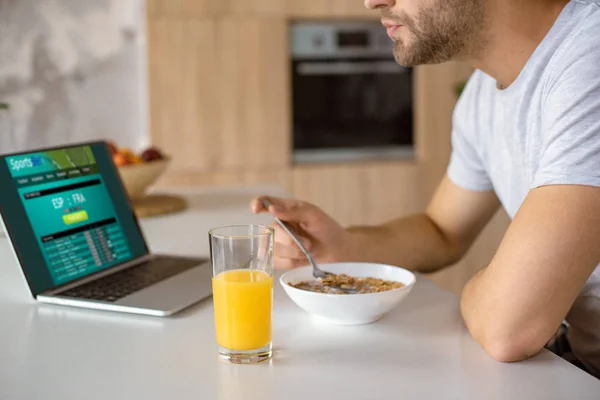 Image recadrée de l'homme mangeant des flocons avec du lait à la table de cuisine avec du jus frais en verre et ordinateur portable avec pari sportif à l'écran — Photo de stock