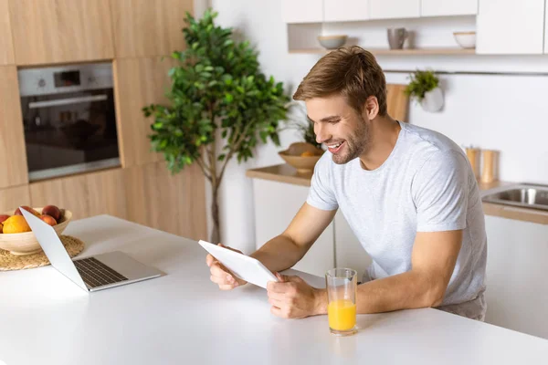 Jeune homme souriant utilisant une tablette numérique à la table de cuisine avec jus et ordinateur portable — Photo de stock