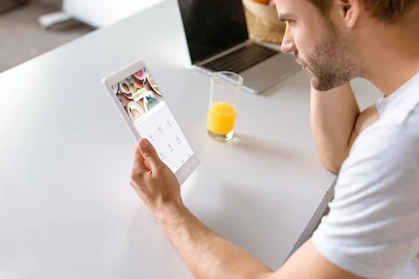 Joven en la mesa de la cocina con el jugo y el ordenador portátil utilizando tableta digital con cuadrado en la pantalla - foto de stock