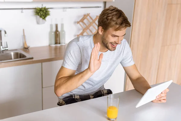 Sorridente giovane uomo agitando da e e parlando da tablet digitale in cucina — Foto stock