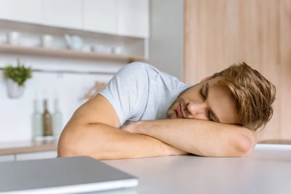 Young man sleeping at kitchen table with laptop — Stock Photo