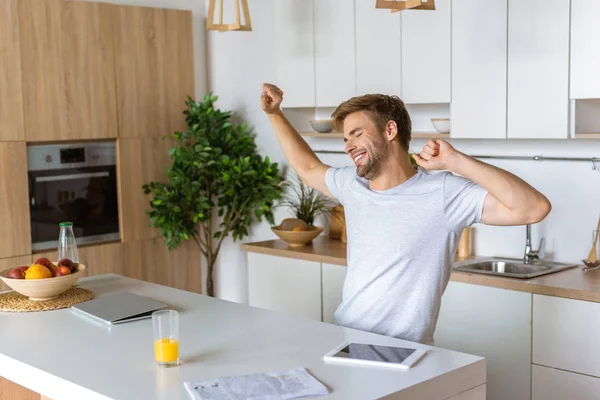 Lächelnder junger Mann mit breiten Armen sitzt am Küchentisch mit digitalen Geräten — Stockfoto