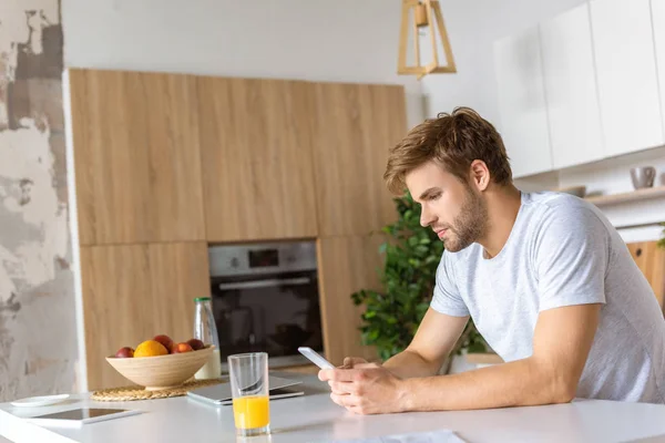 Jovem sério usando smartphone na mesa da cozinha com suco e laptop — Fotografia de Stock