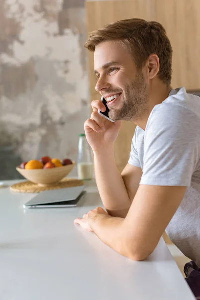 Selective focus of happy young man talking on smartphone at kitchen table — Stock Photo