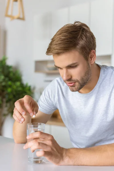 Junger Mann steckt Pille in Glas mit Wasser am Küchentisch — Stockfoto