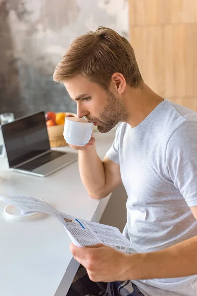 Concentrado joven bebiendo café y leyendo el periódico en la cocina - foto de stock