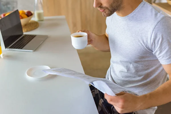 Cropped image of man drinking coffee and reading newspaper at kitchen table with laptop — Stock Photo