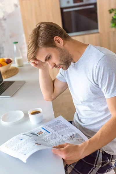 Сосредоточенный человек, читающий газету за кухонным столом с кофе и ноутбуком — стоковое фото