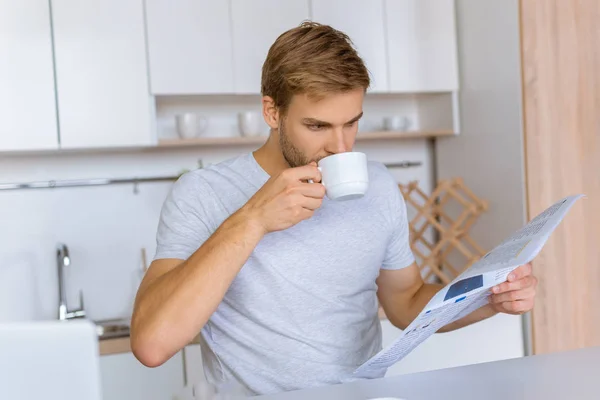 Focused young man drinking coffee and reading newspaper at kitchen table — Stock Photo