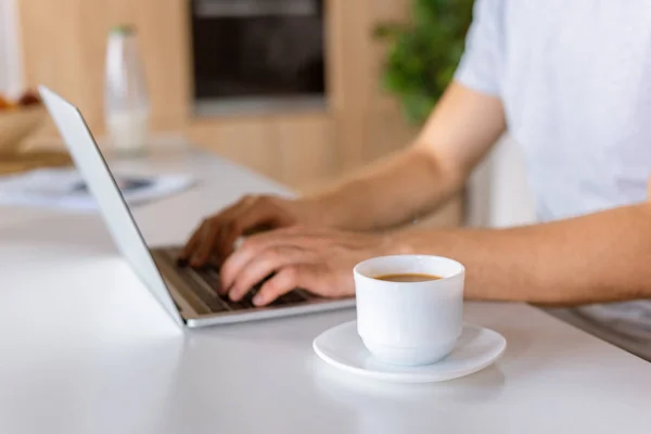 Enfoque selectivo de la taza de café y el hombre usando el ordenador portátil en la mesa de la cocina - foto de stock