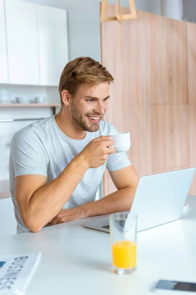 Feliz joven bebiendo café y mirando la pantalla del ordenador portátil en la cocina - foto de stock