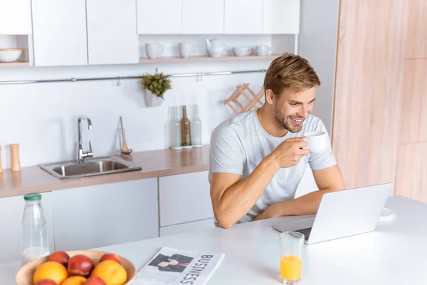 Smiing young man drinking coffee and looking at laptop screen at kitchen — Stock Photo