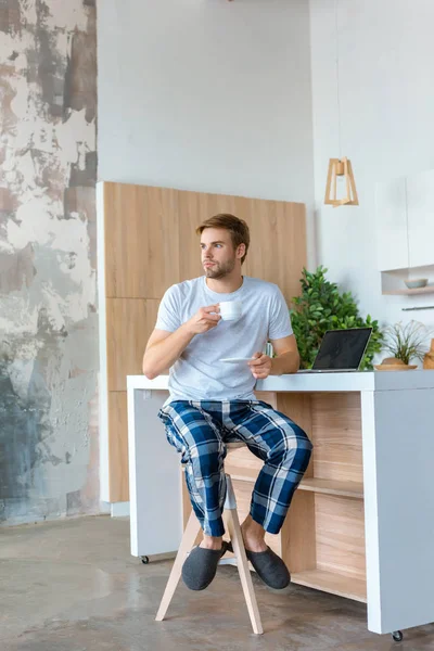 Joven bebiendo café y mirando a la mesa con el ordenador portátil - foto de stock