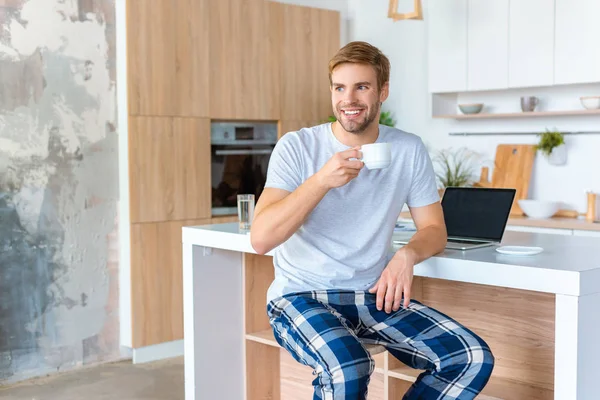 Jeune homme souriant buvant du café à la table de cuisine avec ordinateur portable — Photo de stock